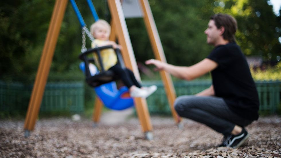 Föräldraledig pappa gungar med sitt barn i en lekpark.