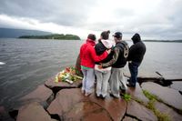 En grupp ungdomar sörjer vid en minnesplats på fast mark. Ute på sjön syns ön Utøya, där polisen fortfarande letar efter saknade.