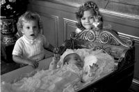 Prinsessan Madeleine i dopklänning. Ligger i ornerad vagga. Victoria och Carl Philip står bredvid sin lillasyster.